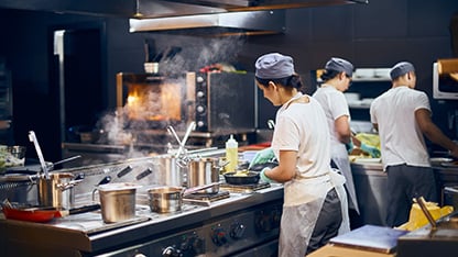 En kvinnlig kock står i ett restaurangkök och tillagar något på spisen. En manlig och en kvinnlig kock jobbar i bakgrunden.